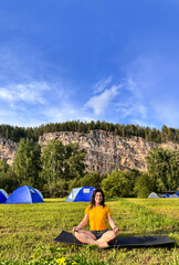 Positive woman meditating in lotus pose at mountain campsite with tents and blue sky, free space, relaxed woman doing yoga outside at campsite with clear sky and copy space, vertical shot