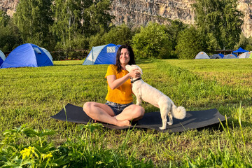 A positive woman in a yellow T-shirt does yoga at a campsite with a white fluffy dog under a blue sky, yoga with dog, outdoor sports, horizontal shot