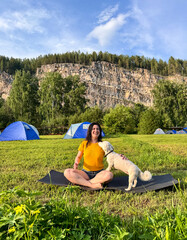 A positive woman in a yellow T-shirt does yoga at a campsite with a white fluffy dog under a blue sky, yoga with dog, outdoor sports
