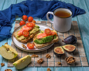 Healthy breakfast on rustic wood featuring avocado toast with salmon and cherry tomatoes, figs, walnuts, apple slices, and herbal tea in soft natural light
