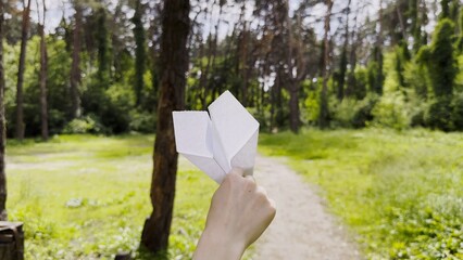 Female hand playing with paper airplane at summer forest. Girl walking with toy plane at woodland on sunny day. Young woman having fun at nature. Concept of leisure outdoor. Close up
