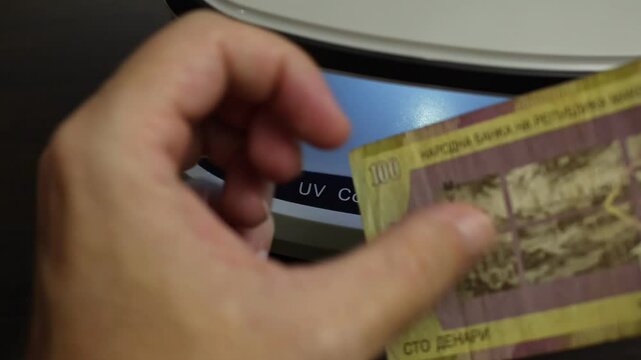 Examination of Macedonian denar cash money with UV counterfeit detector. Concept of authenticity check, fight against counterfeiting of banknotes, anti fraud measures in North Macedonia. Close up view
