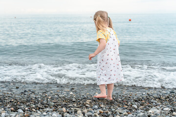 Cute little child in a dress on the beach with rocks on the seashore