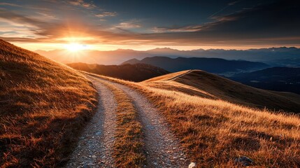 Naklejka premium A winding dirt path crosses golden grassy hills at sunset, with distant mountain ranges under a vibrant sky.