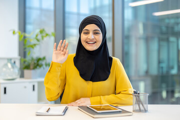 A smiling woman in a hijab waves at the camera, looking ready for a video call