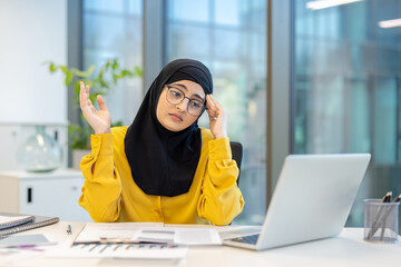 A woman wearing a hijab looks stressed while working on a laptop in a modern office setting.