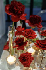 Vibrant red roses and peonies arranged gracefully in glass vases on a table. Candles placed around...
