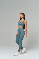 Fit woman in blue athletic wear and white sneakers stands confidently on one leg, showcasing toned physique, long hair, and neutral expression against a minimalistic setting