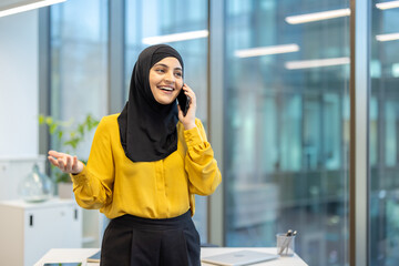 A cheerful woman wearing a hijab speaks on the phone in a bright office setting.
