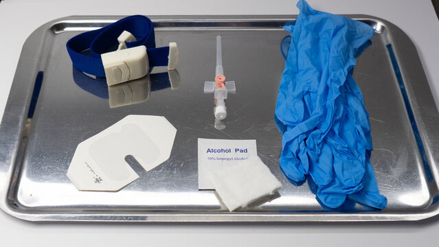 Sterile IV cannulation kit arranged on a stainless steel tray, including gloves, pink needle cannula, alcohol pad, gauze, tourniquet, and transparent IV dressing.