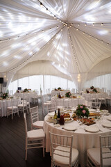 Elegant indoor event setting featuring round tables with white linens and arranged floral centerpieces. Chairs are neatly arranged, and the ambiance suggests a formal celebration or gathering.