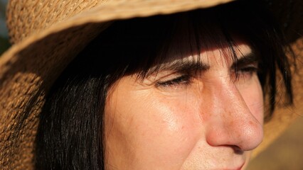 Female farmer in straw hat looking into wheat field and examining plantation at sunset. Close up face of adult beautiful woman illuminating by bright rays of sun. Concept of agriculture business