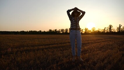 Adult agronomist in straw hat exploring barley on meadow at sunset. Female farmer examining ripe wheat at field on dusk. Beautiful scenic landscape. Concept of agriculture and agronomy business