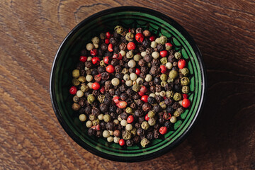 Colorful peppercorn mix in green bowl. Spices on wooden table background. Natural culinary seasoning ingredients. Aromatic pepper grains in round ceramic dish. Flavorful rustic cooking concept.