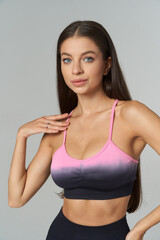 Young woman with long dark hair wears a pink and black sports bra, smiling slightly with one hand on her collarbone, showcasing fitness attire and healthy lifestyle