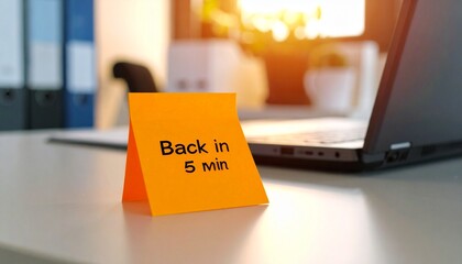 Orange sticky note reading "Back in 5 min" on a desk with a laptop nearby