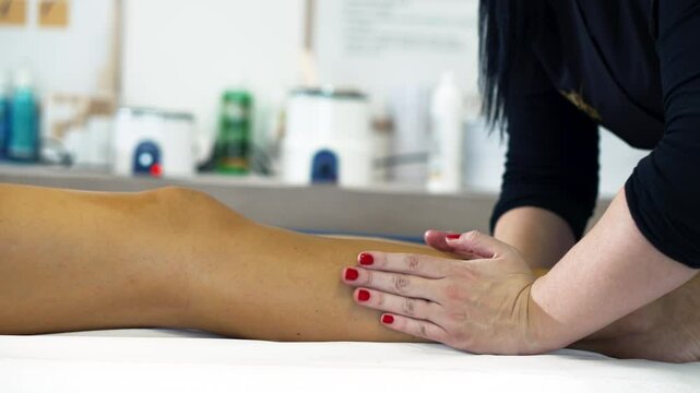 A depilatory specialist in a black uniform uses massaging movements of her palms to relieve skin irritation after hair removal on women's tanned shins in beauty salon. Beauty services in the spa salon