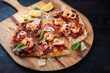 Traditional Italian pinsa bread prosciutto e salsa di pomodoro with king prawns, squid rings and parmesan cheese served with tomatoes and sauce as close-up on a wooden pizza shovel