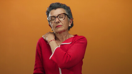 Senior woman in red shirt with grey hair and glasses experiences shoulder pain against an orange...