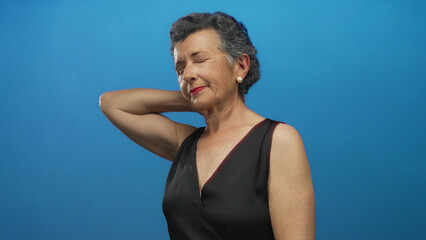 Senior woman with grey hair in black dress against blue background, expressing neck pain, with...