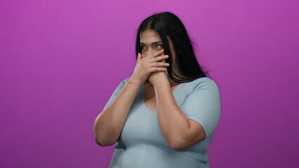 Woman covers mouth with hands standing against pink background, expressing surprise, wearing blue...
