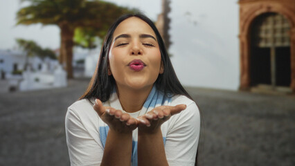 Woman purses lips in blowing pose within outdoor scene by a young latin hispanic figure, delivering...