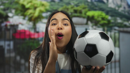 Young hispanic woman outside celebrating with a soccer ball in an urban street environment, showing...