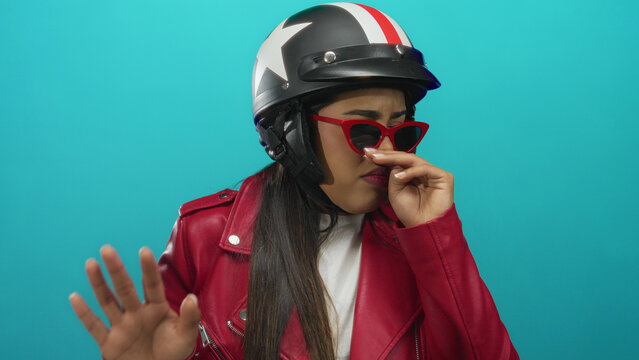 Woman holds nose to pinch foul air near a wall while wearing young hispanic helmet and standing by motorbike against a green isolated backdrop. - Powered by Adobe