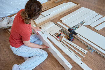 Woman assembling white furniture parts on wooden floor using drill, work tools and instruction manual. Concept shows DIY home improvement, furniture assembly and home renovation