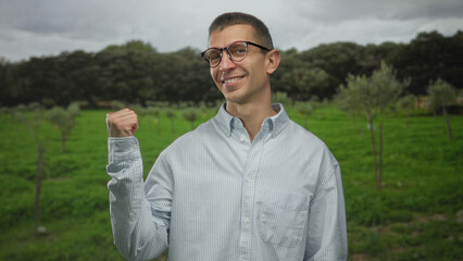 Man smiling with thumbs pointing back in a vibrant outdoor park setting, wearing glasses and a...