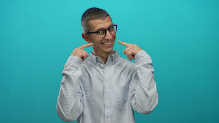 Fototapeta premium Man in glasses and striped shirt pointing at his smile against a blue background, exuding confidence and joy in an isolated studio setting.