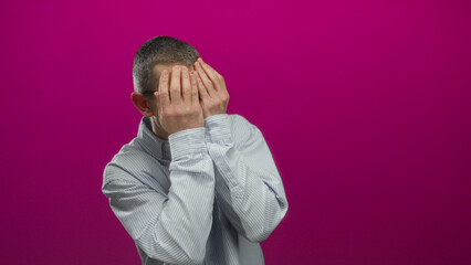 Man making funny faces with hands over eyes against a bright pink background, wearing striped shirt...