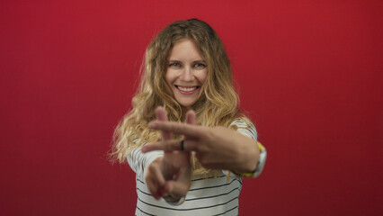 Woman holds hands in hashtag gesture inside studio where blonde young subject poses against red wall with smile wearing watch and striped top.