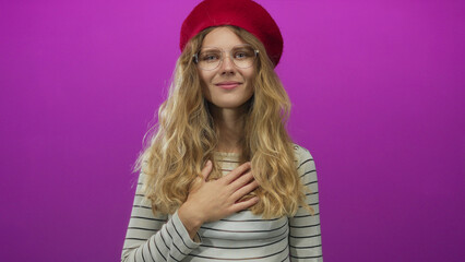 Fototapeta premium Woman gently places a hand over her heart in gratitude inside a studio as a blonde young model wearing glasses and a red beret poses in a striped shirt against a purple backdrop.