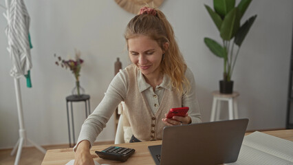 Woman balances phone while calculating budgets in a bright office with a blonde young home setup...
