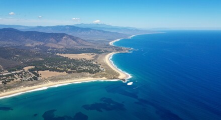 Aerial view of a pristine coastline, showcasing a long sandy beach meeting the ocean, backed by mountains under a clear blue sky
