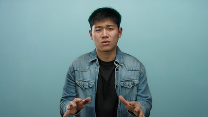 Young chinese man wearing denim jacket gestures expressively against isolated blue background...