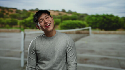 Young man smiling outdoors on tennis court with greenery in the background, capturing a joyful...