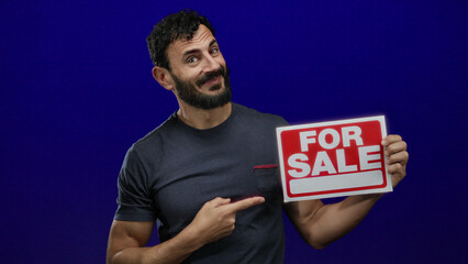 Hispanic man with beard holding a for sale sign against a blue background, pointing confidently...