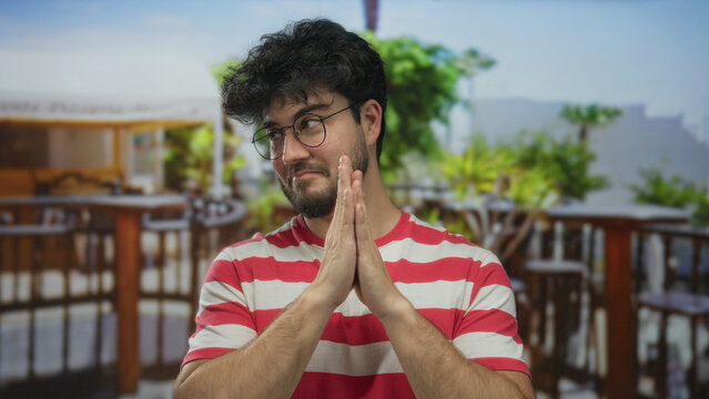 Young man with beard and glasses in red striped shirt stands with joined hands on an outdoor restaurant terrace with lush greenery and wooden railings.