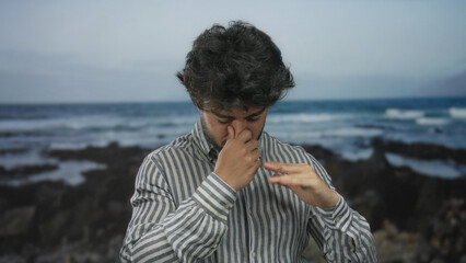 Young man with beard holding nose outdoors at the seaside due to bad smell while standing by the...