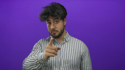 Young hispanic man with beard pointing forward and making a call gesture in front of purple background, wearing striped shirt, conveying communication and engagement.