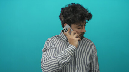 Young man with beard talking on smartphone against blue background wearing casual striped shirt looking thoughtful and focused on a call.