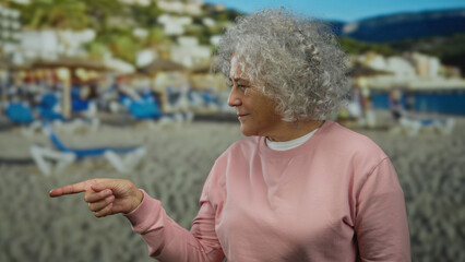 Woman with grey hair in pink sweatshirt points at beach background with blue sunbeds and umbrellas,...