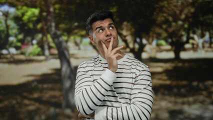 Young man with thoughtful expression in a striped shirt contemplating in a sunny park setting surrounded by trees showcasing a serene outdoor environment.