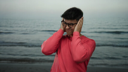 Man on beach with eyes closed covering ears wearing glasses and red shirt by the sea under overcast...