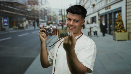 Young man holding vintage camera gestures invitingly on busy city street during daytime in outdoor...