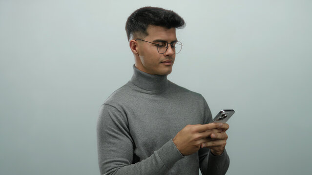 Young man with glasses holding smartphone against a plain background, displaying a focused expression.