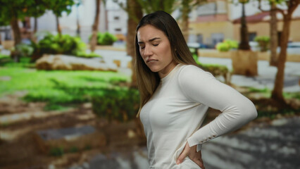 Young woman with back pain stands in an outdoor park setting, expressing discomfort amidst vibrant...