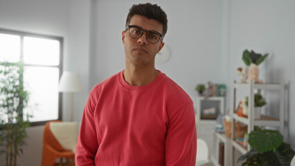 Young man in glasses and red sweater stands thoughtfully in a cozy bedroom adorned with plants and...